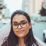 A confident woman wearing glasses smiles subtly while standing on an urban street with blurred background.